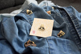 An A enamel pin with butterfly backer delicately handlettered in black, white, and gold on a floral board backer on top of a jean jacket surrounded by other enamel pins