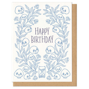 Greeting card with kraft paper envelope. "Happy birthday" hand written in serif font, centered. Surrounded by light blue leaves and skulls with hourglass at bottom.