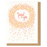 white greeting card with an illustrated circle of yellow and orange confetti surrounded hand-lettering that reads "so proud of you"
