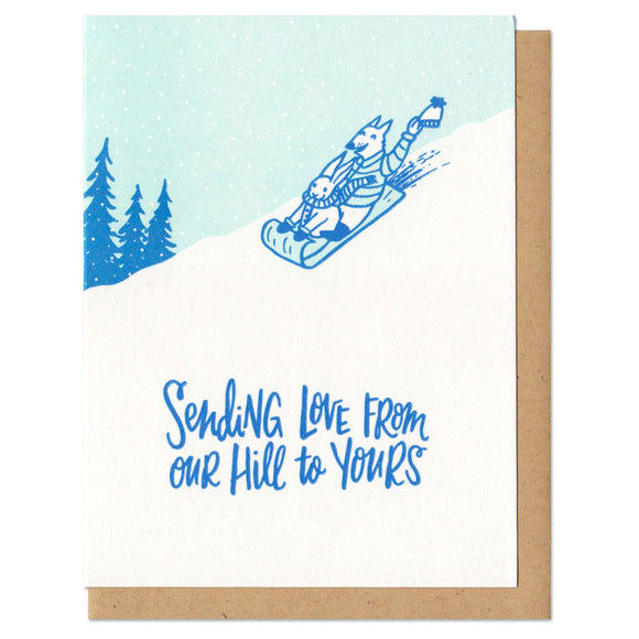 white greeting card with an illustration of a wolf and rabbit dressed for winter, sledding down a hill by silhouetted pine trees. blue hand-lettering beneath them reads 