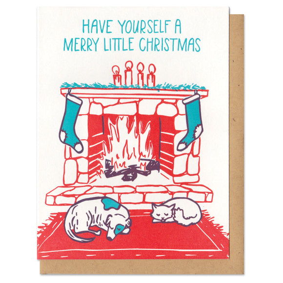 A greeting card and envelope featuring an illustration in green and red of a fire place with stockings hanging and a cat and dog laying in front of it. The text abouve reads 
