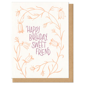 A greeting card and envelope that reads "happy birthday, sweet friend" in light purple with orange flowers surrounding the text.