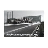 black and white postcard photograph of night-time traffic driving over a downtown providence bridge with the iconic power station smoke stacks visable in the background. white text on the bottom reads "greetings from providence, rhode island"