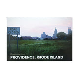 postcard photograph of the Providence skyline with a "road closed" street sign in the foreground. white text on the bottom reads "greetings from providence, rhode island"