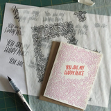 greeting card with pink leaf pattern and red text which reads "you are my happy place" pictured with design sketches and a pen