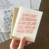 hand holding a white greeting card with red text which reads "you are a fierce lady-dragon who breathes fire upon trolls, hater, and mansplainers" above design sketches