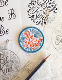 A circular iron on patch with blue floral and a coral script that reads "be kind".
