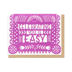 Greeting card and kraft paper envelope. Card reads "celebrating you is easy" in illustrated papel picado style cut paper flag in purple