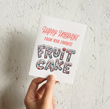 A greeting card and envelope featuring script that says "Happy holidays from your favorite" with "fruit cake" written below in the shape of an actual fruitcake.