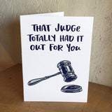 white greeting card with navy text that reads "that judge totally had it out for you" above an illustration of a gavel