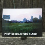 postcard photograph of the Providence skyline with a "road closed" street sign in the foreground. white text on the bottom reads "greetings from providence, rhode island"
