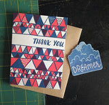 white greeting card with a red and blue illustrated triangles pattern and hand lettering that reads "thank you" photographed with a blue could "dreamer" patch