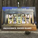 postcard featuring a photograph of the iconic Arcade mall lit up in the evening. white text reads "Providence, RI" photographed in front of a fence