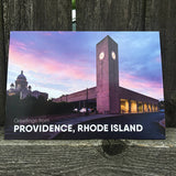 postcard photograph of the providence train station at dusk with the state house visable in the background. white text at the bottom reads "greetings from providence, rhode island"