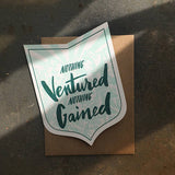 badge-shaped greeting card with a light blue illustration of a mountain range behind green hand-lettering that reads "nothing ventured nothing gained" photographed in cast natural light