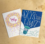 white greeting card featuring an illustration of two penguins riding in a hot air balloon. hand-lettering on the balloon reads "sky's the limit" pictured with a "so proud of you" greeting card