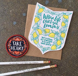 Die cut badge-shaped greeting card that reads "when life give you lemons, try to figure out something to do with those lemons" in teal text surrounded by illuatrated lemons and leaves photographed with a "take heart" patch and two white pencils