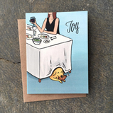 A greeting card with an illustration of a dinner table with a yellow dog under it who is eating a chicken leg with the word Joy above.