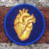 An round embroidered patch with an iron on back featuring a golden anatomical heart with a navy blue background. 
