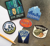 Six patches, from left to right: a blue book smeller, clouds that say dreamer, a red oval that says chasing wonder, three pigeons with text that says wild in the street, a flashlight illuminating mystery solver, and a hermit crab with wanderer.