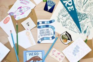 A collection of greeting cards, stickers, prints, patches, and pencils featuring board games, a hermit crab, flowers, pangolins, and a rocket. The pencils feature lyrics from popular hip hop songs.