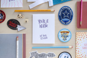 A letterpressed greeting card that says, "you're a weird, but you're my weird" surrounded by a cold pizza club patch, a glow in the dark stargazer patch, and a few pencils.