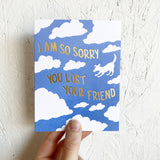 blue greeting card with a white cloud pattern featuring the silhouette of a dog. gold foil stamped lettering reads "i am so sorry you lost your friend" hand-held in front of a white wall