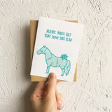 Greeting card and kraft paper envelope. Teal, hand-written text that reads, "Maybe you'll get that pony this year." Illustration of pony wrapped in wrapping paper with bow below text. Shades of teal. PHotographed hand-held in front of a white wall.