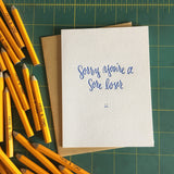 white greeting card with blue text that reads "sorry you're a sore loser" above a small frowny face drawing photographed with pencils