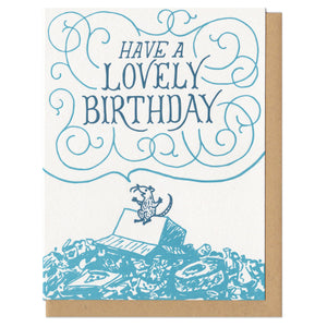 Greeting card and kraft paper envelope. Illustration of rat on top of garbage pile happily saying "have a lovely birthday" in hand-written delicate font surrounded by swirls. Drawn in shades of blue.