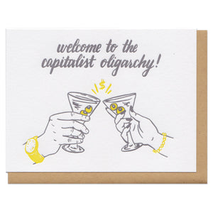 white greeting card with an illustration of two hands cheers-ing martini glasses beneath grey text that reads "welcome to the capitalist oligarchy!"