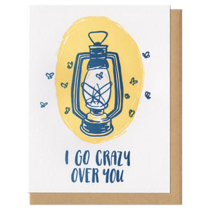 Greeting card with kraft paper envelope. Illustration of old camping oil lamp with moths flying around it. Handwritten text below it reads, "I go crazy over you."