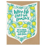 Die cut badge-shaped greeting card that reads "when life give you lemons, try to figure out something to do with those lemons" in teal text surrounded by illuatrated lemons and leaves