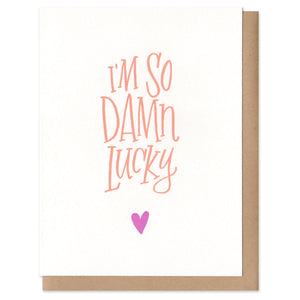Greeting card and kraft paper envelope. Handwritten text in orange reads, "I'm so damn lucky." Tiny pink hear below it.