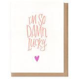 Greeting card and kraft paper envelope. Handwritten text in orange reads, "I'm so damn lucky." Tiny pink hear below it.