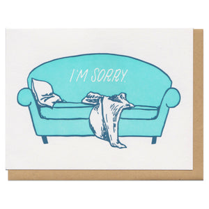 white greeting card featuring an illustrated teal couch with a pillow and blanket slumped on it. white text on the couch reads "I'm sorry"