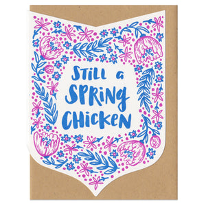 Die-cut, shield shaped greeting card with kraft paper envelope. Handwritten type in navy reads, "Still a spring chicken." Surrounded by navy and purple flowers and leaves.