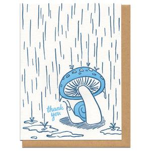 white greeting card witn an illustration of a snail hiding from the rain beneath a mushroom next to hand-lettering that reads "thank you"