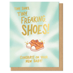 Greeting card with kraft envelope that reads, "tiny shoes. tiny. freaking. shoes! congrats on your new baby!" Letters are in gold foil. Illustration of tiny converse-like baby shoes in peach with light radiating from them. 