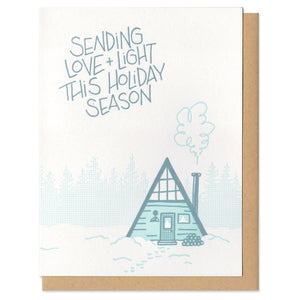 white greeting card with a blue and teal illustration of an a-frame cabin and tree line of pines. hand lettering above the house reads "sending love + light this holiday season"