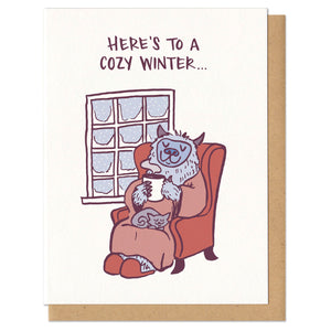 A greeting card and envelope featuring capitalized text that reads, "here's to a cozy winter..." with a yeti bundled up in a robe and slippers, with a mug in hand and cat on their lap in front of a snowy window. 
