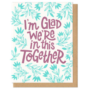 A greeting card and envelope featuring mint green buds and stems with hand lettered text in the center printed in purple that reads "I'm glad we're in this together"