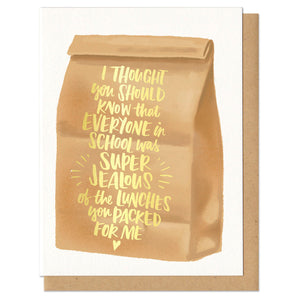 Greeting card and kraft paper envelope. Illustration of brown paper bag with text on it in gold foil that reads, "I thought you should know that everyone in school was super jealous of the lunches you packed for me."