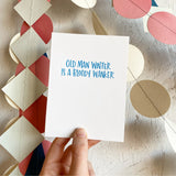 white greeting card with blue hand-lettering that reads "old man winter is a bloody wanker" being hand-held in front of colorful paper garlands