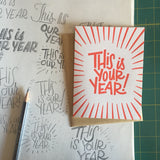 white greeting card with red lines and hand-lettering that reads "this is your year!" photographed with design sketches