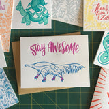 white greeting card with an illustrated ant-eater on rollerskates beneath hand-lettering that reads "stay awesome" photographed amongst other frog & toad press cards