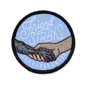 A circle iron on embroidered patch with a black outline and a light blue background with a dog's paw holding a human's hand with scripted text above it reading "friend to strays"