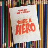 white greeting card with red text which reads "you're more than a teacher - you're a hero" shown atop a pile of pencils