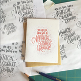 white greeting card with red hand lettering that reads "you are like some sort of mythical godess creature" pictured with designing sketches and tools
