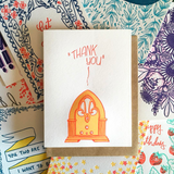 white greeting card with an illustrated old fashioned radio, printed in orange, beath hand-lettering that reads "thank you" photographed on top of an assorment of other Frog & Toad Press greeting cards